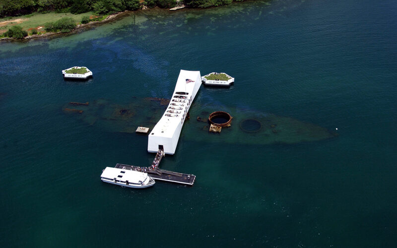 Aerial view of the USS Arizona Memorial at Pearl Harbor with the sunken battleship visible beneath the water and a tour boat docked alongside