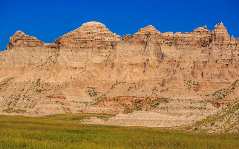 Dramatic layered rock formations rising above green prairie grass under blue sky at Badlands National Park in South Dakota